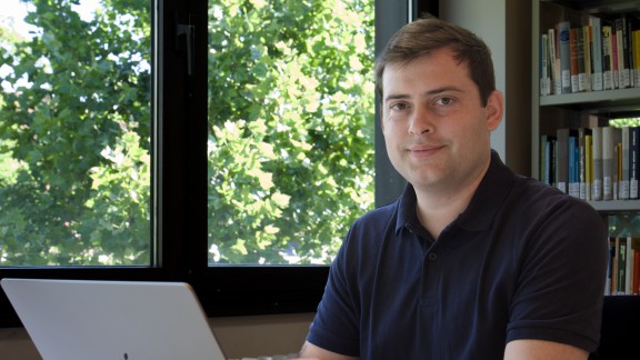 Foto zeigt Wenzel Massag (links) vor einem Bücherregal und einem Fenster durch das grüne Bäume zu sehen sind.
